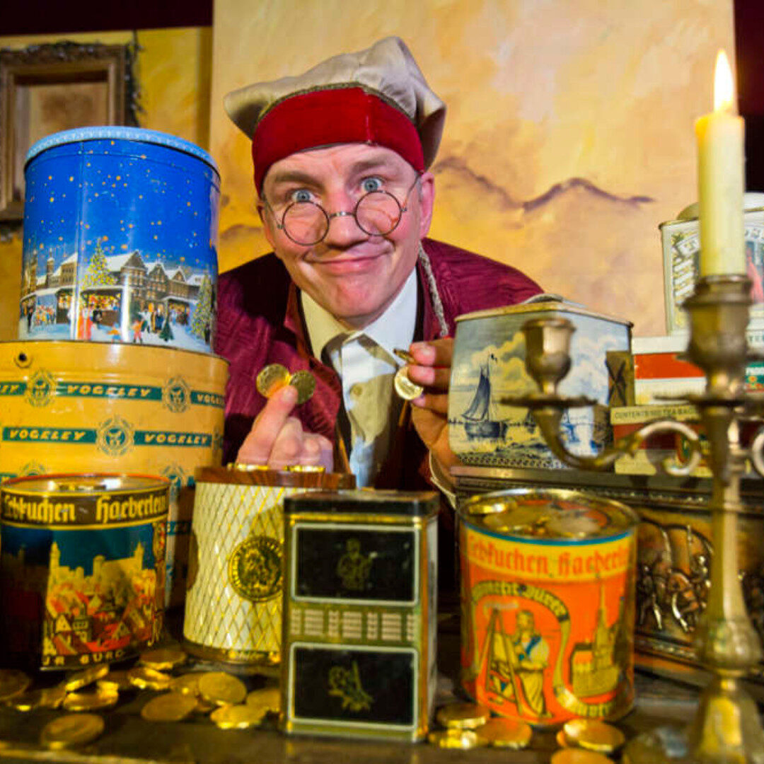 Foto aus dem Kindertheater William Goldtaler der Zweite!. Ein Mann mit Goldmünzen in der Hand und verschiedenen Dosen vor ihm auf dem Tisch.